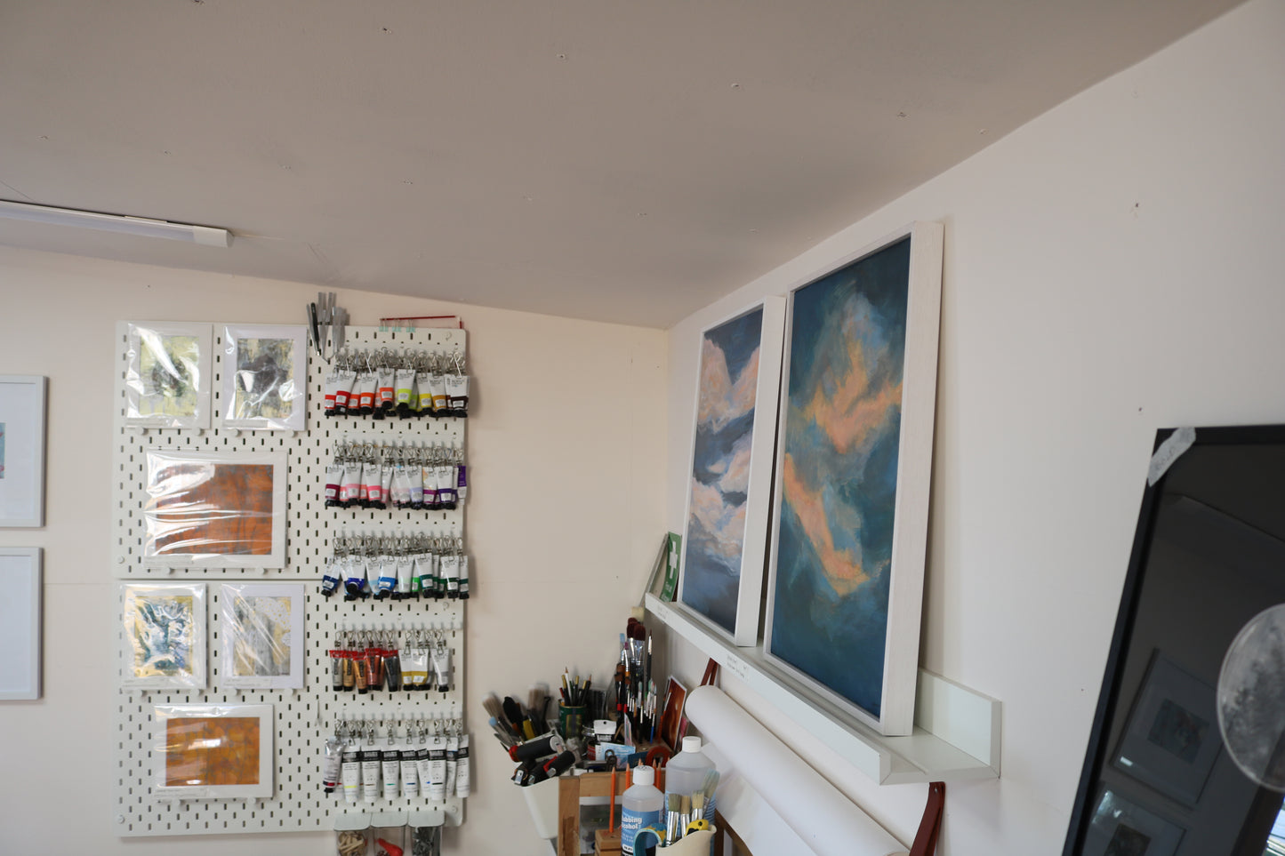 Image shows Autumn Dawn 2 painting from the right hand side, taken at an angle. It sits alongside another of Kate's sky paintings on to of a white shelf. Other aspects of Kate's studio can also be seen including paintbrushes in pots and acrylic paint tubes hung on a organiser on the wall.