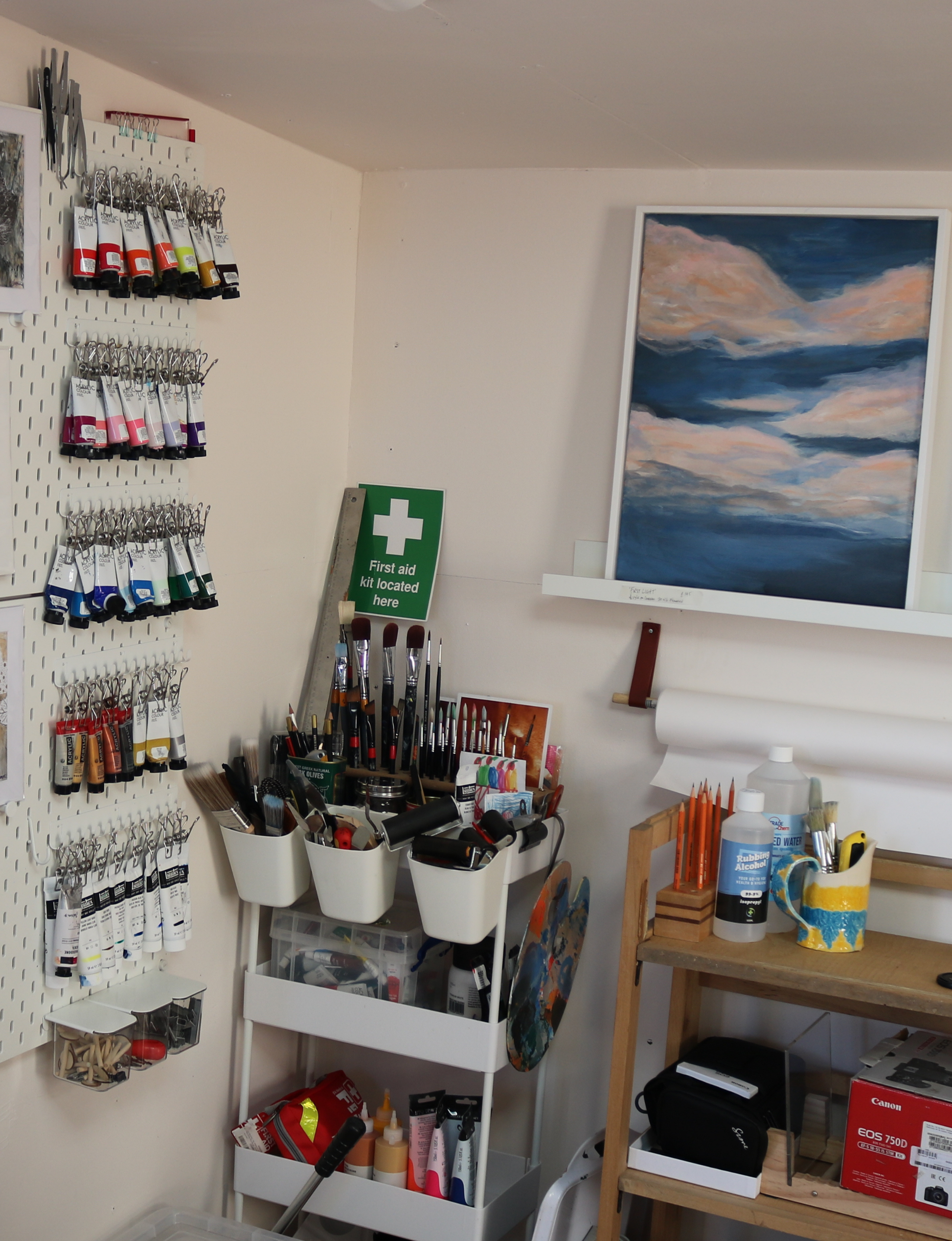 The image shows a corner of the artist's studio. The painting 'First Light' is seen, framed, on a white shelf, below and to the left, in the corner of the room, is a small trolley full of art supplies, paintbrushes and printing brayers. To the left on the adjacent wall, a quantity of acrylic paint tubes are stored on a peg board, attached by metal clips. The walls are a very light gypsum plaster colour.